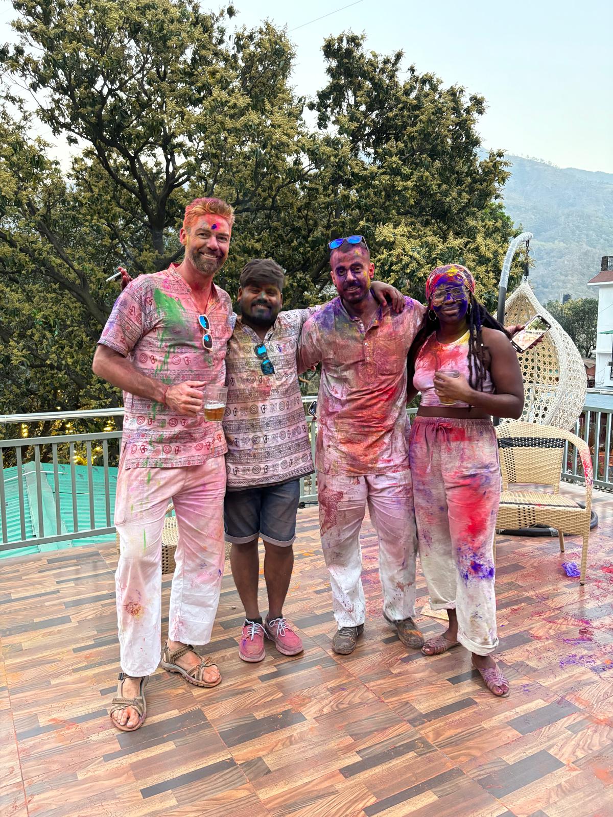 Grupo celebrando Holi, cubiertos de colores en una terraza con vistas montañosas.