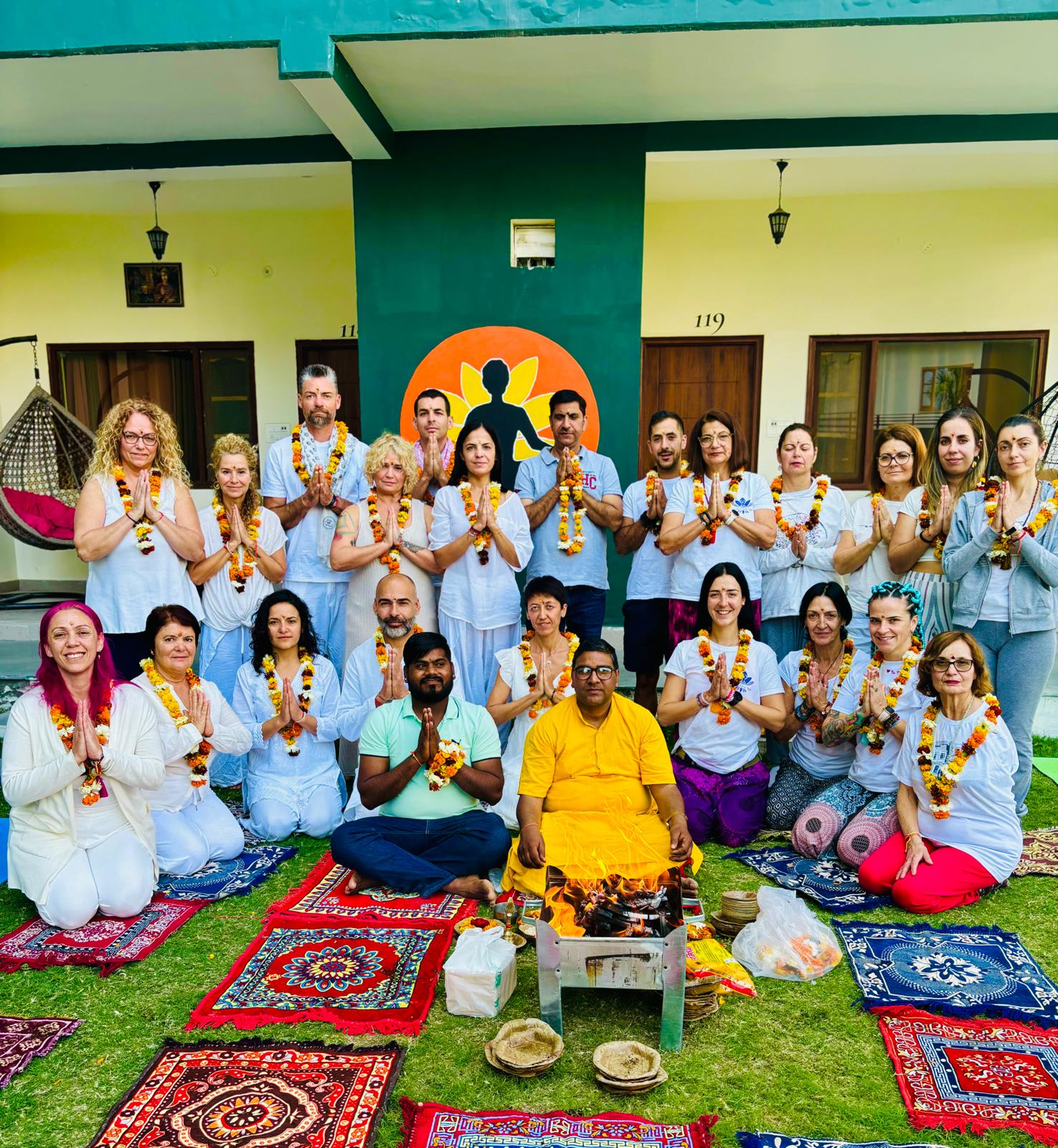 Grupo de viajeros en ceremonia de meditación al aire libre, rodeados de tapetes y flores.