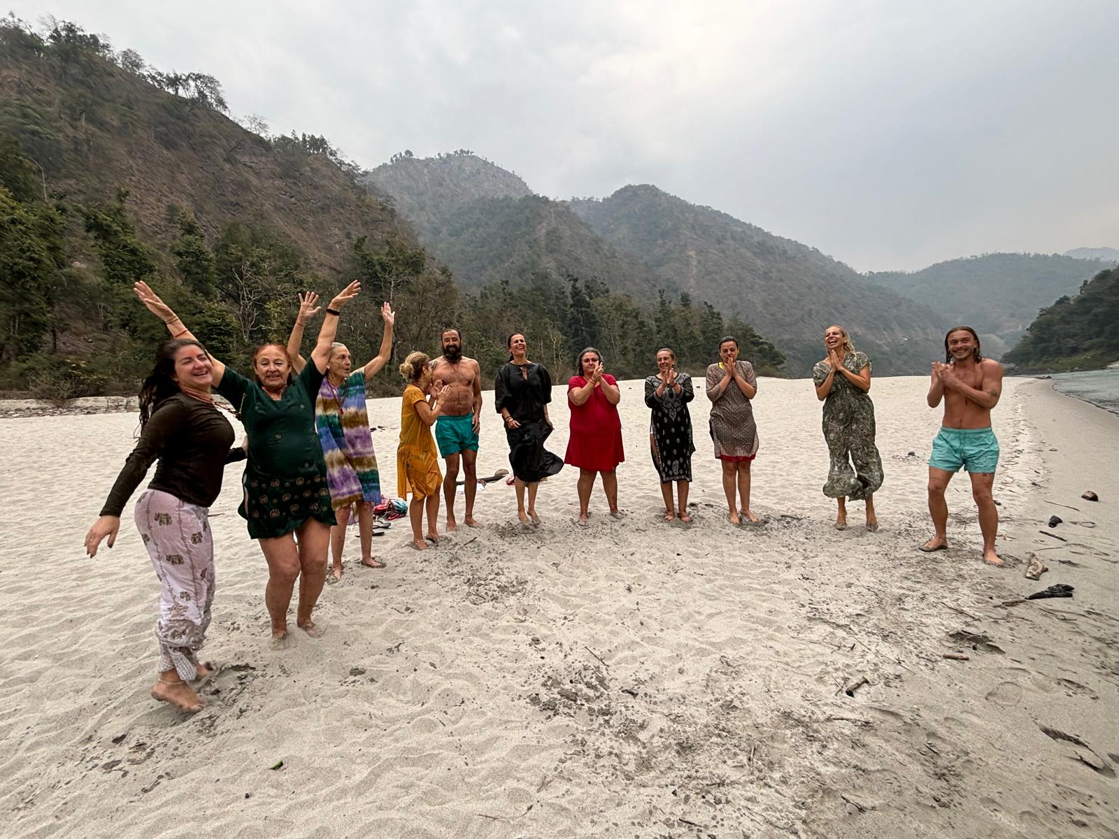 Un grupo de viajeros salta y aplaude en una playa fluvial rodeada de montañas y bosque.